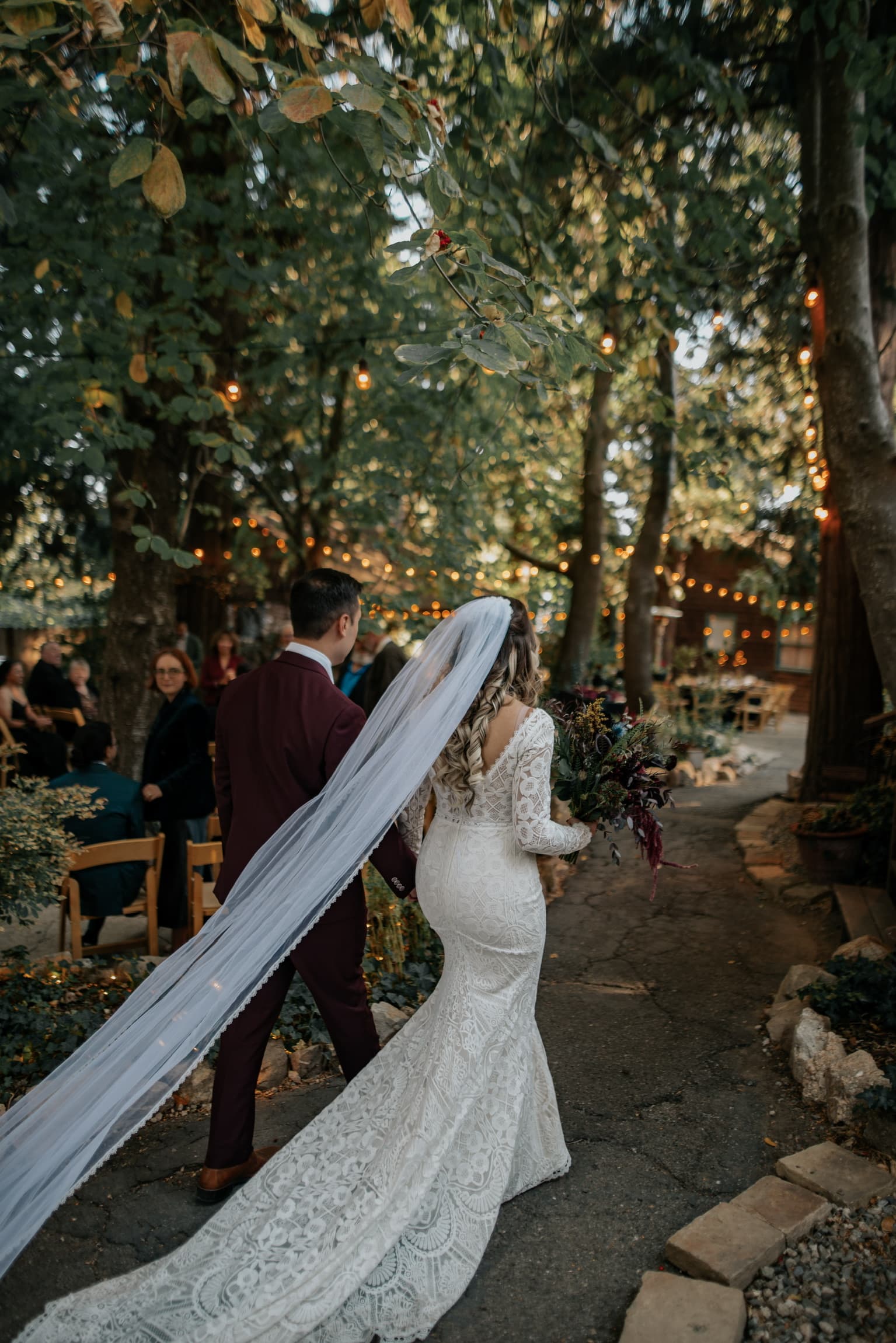 A bride and groom walking back down the aisle through the pines at Willow Woods Park, string lights overhead